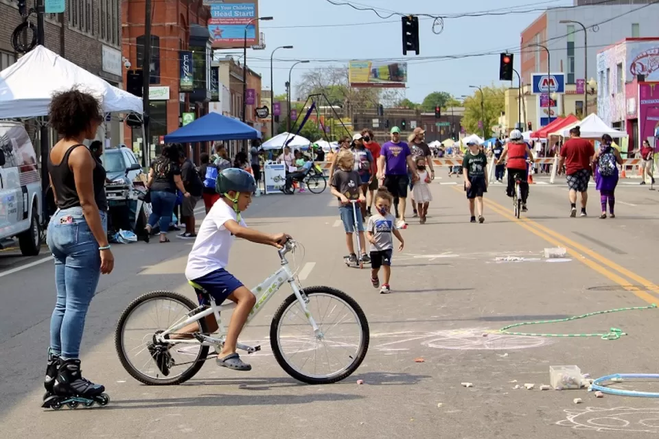 Diverse people of all ages walking and biking at Minneapolis Open Streets event with temporary festival booths along the sides of the street